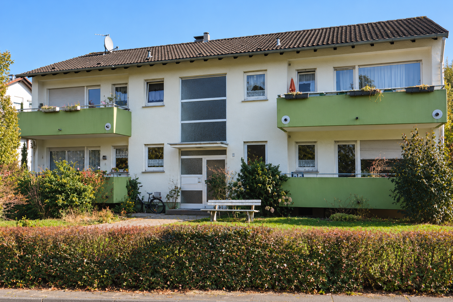 Zweigeschossiges Wohnhaus mit heller Fassade, grünen Balkonen und einer Hecke im Vordergrund 